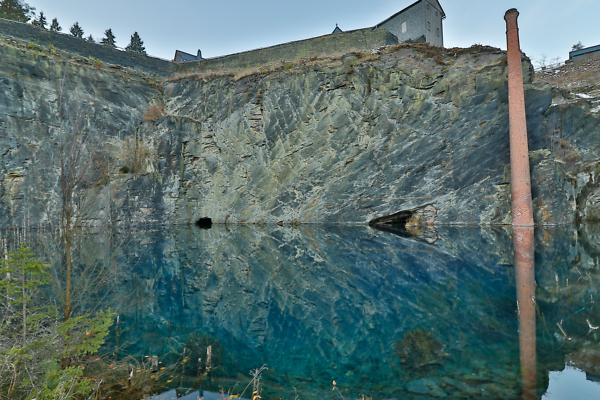 Eine steile Schiefer-Felswand ragt aus dem Wasser auf. Schiefersee am Technischen Denkmal Lehesten.