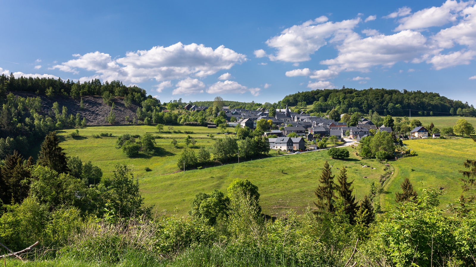 Ein Dorf mit schiefergedeckten Dächern liegt inmitten einer Mittelgebirgslandschaft aus Wiesen und Wäldern.