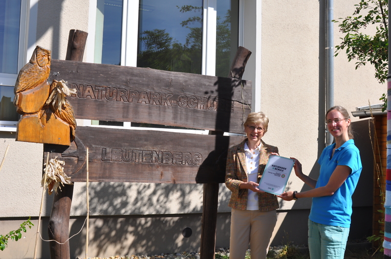 Zwei Frauen stehen vor einem großen Holzschild mit der Aufschrift Naturpark-Schule Leutenberg. Die Mitarbeitende der Naturpark-Verwaltung überreicht der Direktorin der Schule eine Urkunde.