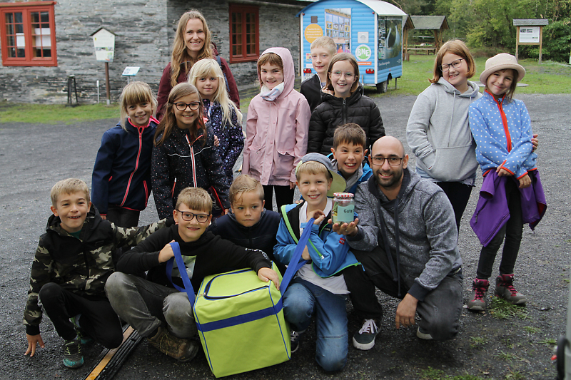 Eine Gruppe Schulkinder und ihr Lehrer lächeln in die Kamera. Im Hintergrund ist das BNE-Mobil am Technischen Denkmal Lehesten zu sehen.