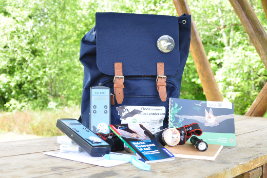 Fledermausrucksack mit samt seinem Inhalt, präsentiert auf einem Holztisch im Freien.